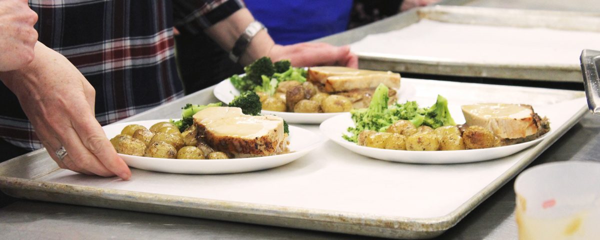 Close up of someone holding a tray bearing three plates of meat, broccoli, and potatoes. A line of other people and trays can be seen out of focus and lined up next to this person.
