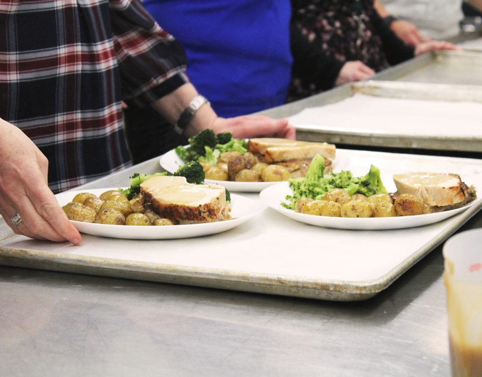 Close up of someone holding a tray bearing three plates of meat, broccoli, and potatoes. A line of other people and trays can be seen out of focus and lined up next to this person.