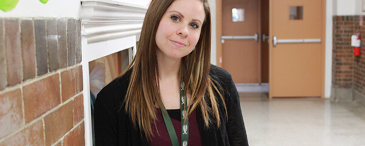 A young white woman with brown hair leans against the wall in a hallway. She smiles at the camera and is wearing a green lanyard.