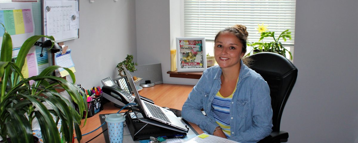 A young woman sits at a desk with plants, a binder, and a telephone. She smiles at the camera.