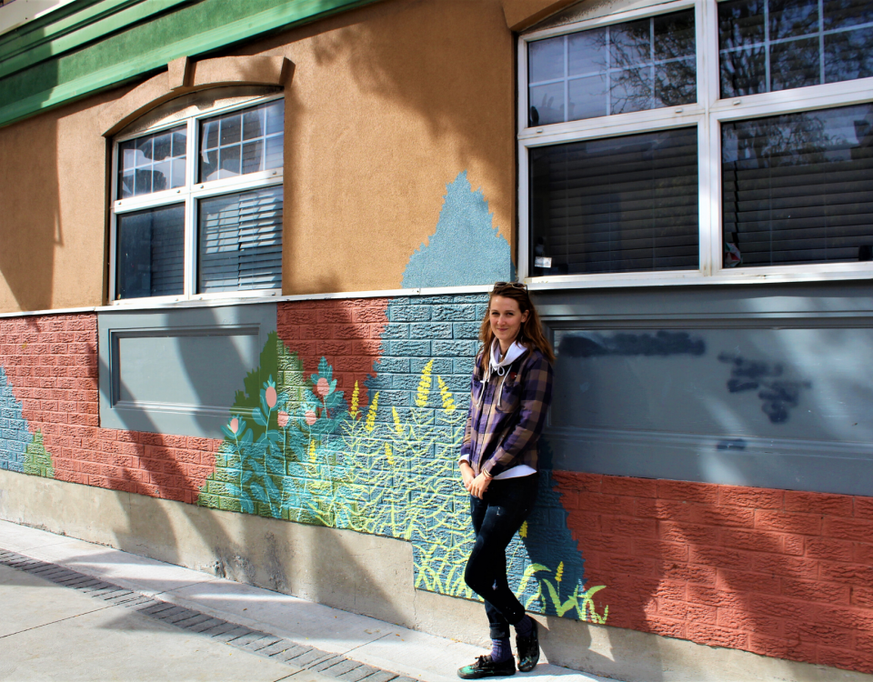 A woman wearing a hooded jacket leans against a brick wall which is painted with a mural of trees and flowers.