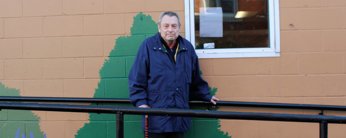 An older man wearing a blue jacket stands on a ramp outside a building, holding a cane in one hand and the railing. His right foot is in a cast. The building has murals of a tree on it.