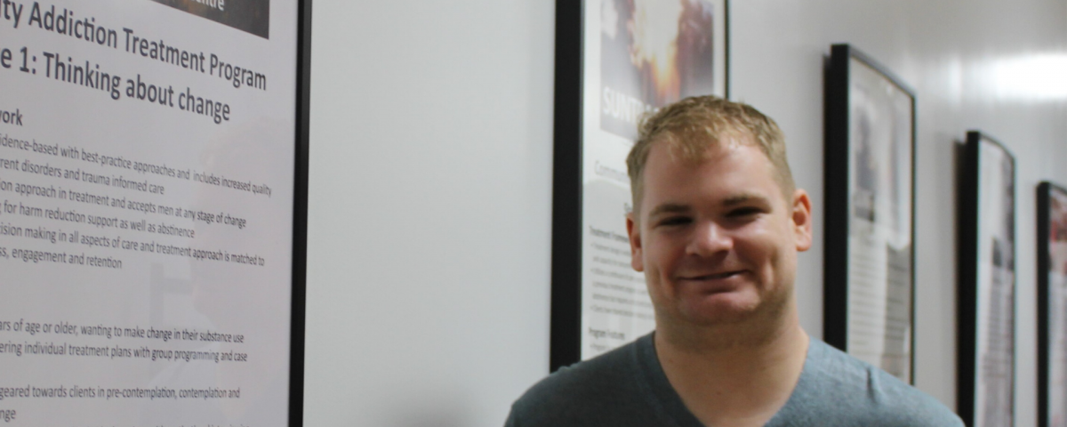 A man with light hair smiles to the camera. He is standing in a hallway with posters describing addiction treatment programs.