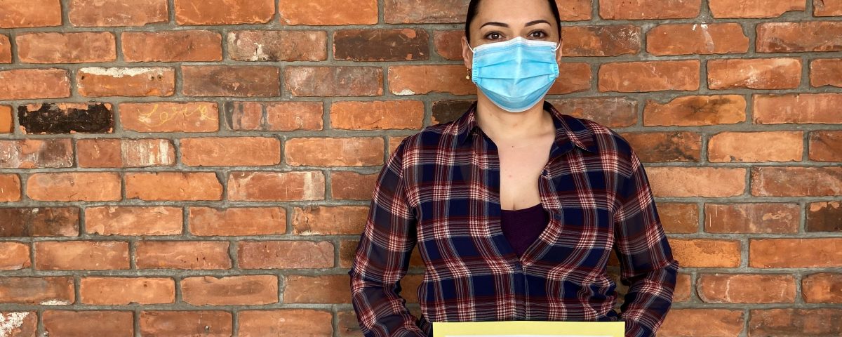 A woman with dark hair pulled back wears a blue surgical mask and a plaid blouse. She stands in front of a red brick wall and holds up a sign that reads "Thank you for helping vulnerable women and children during COVID-19."