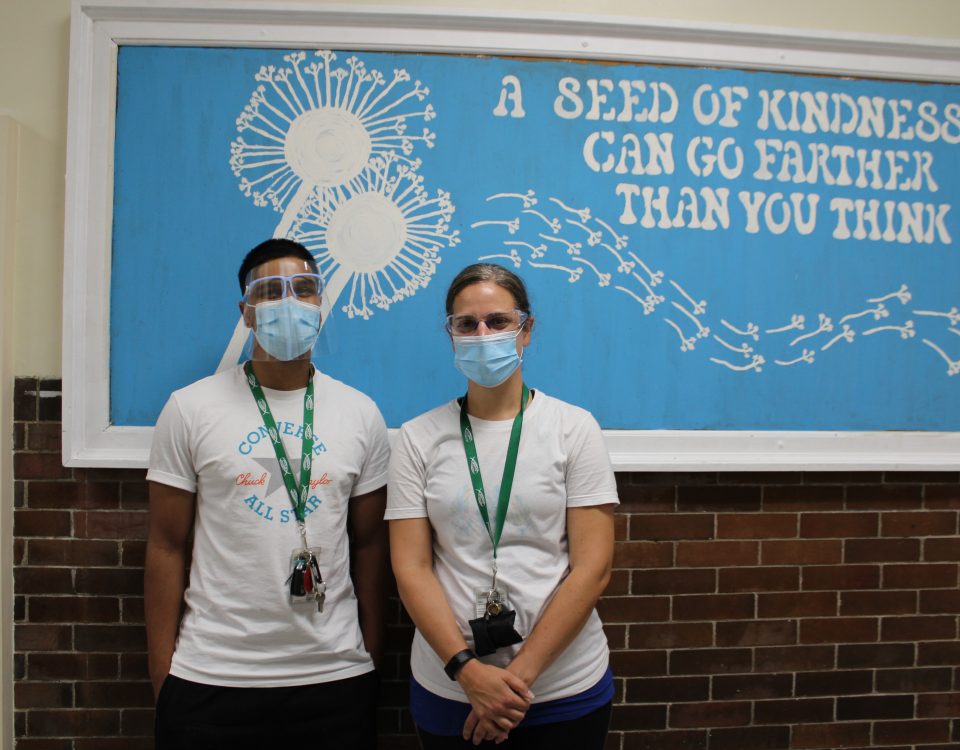 A man and a woman stand in front of a blue bulletin board decorated with a large dandelion and the quote: "A seed of kindness can go further than you think." The two people are wearing white t-shirts, masks, and green landyards.